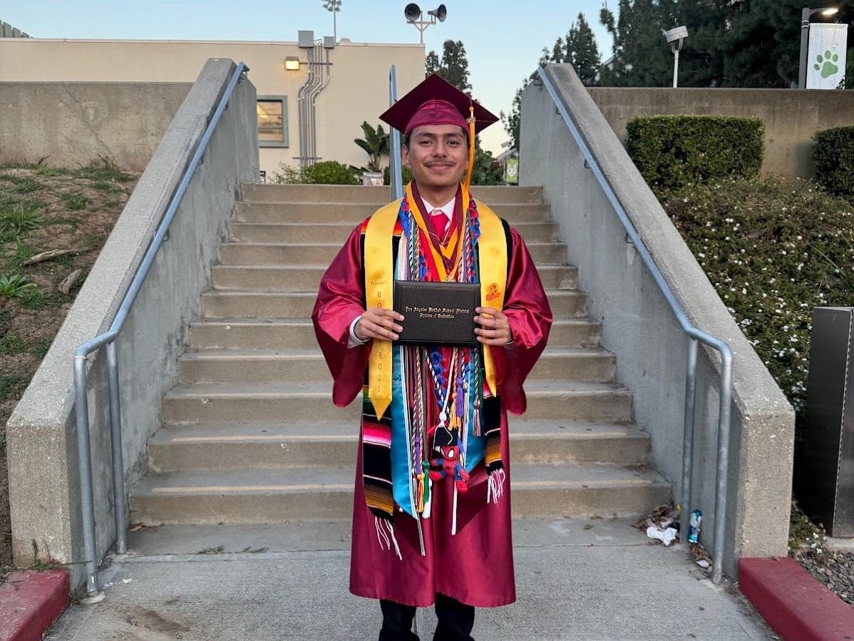 An MFC graduate in a cap and gown holding a diploma on the steps of their school