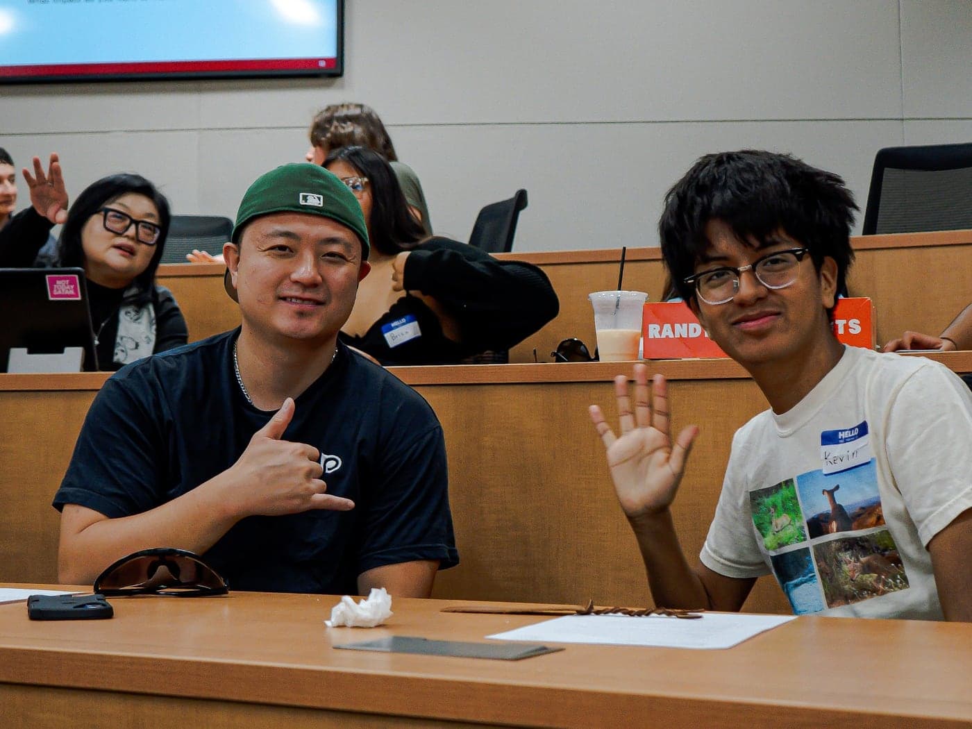 A mentor and mentee smiling and waving together in a campus lecture hall
